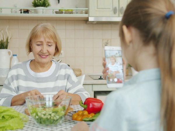 Cheerful elderly woman is posing for smartphone camera and cooking salad while child is taking picture with modern device. Family, photograph and people concept.