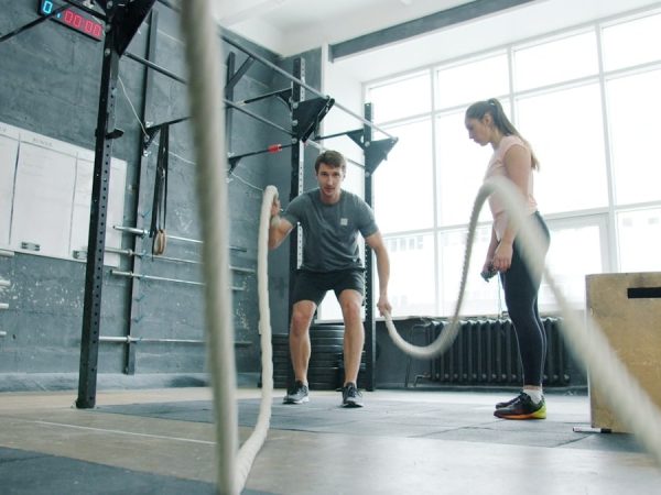 Energetic sportsman is battling ropes working out during sports training with coach young woman in modern gym. Active youth and workout concept.